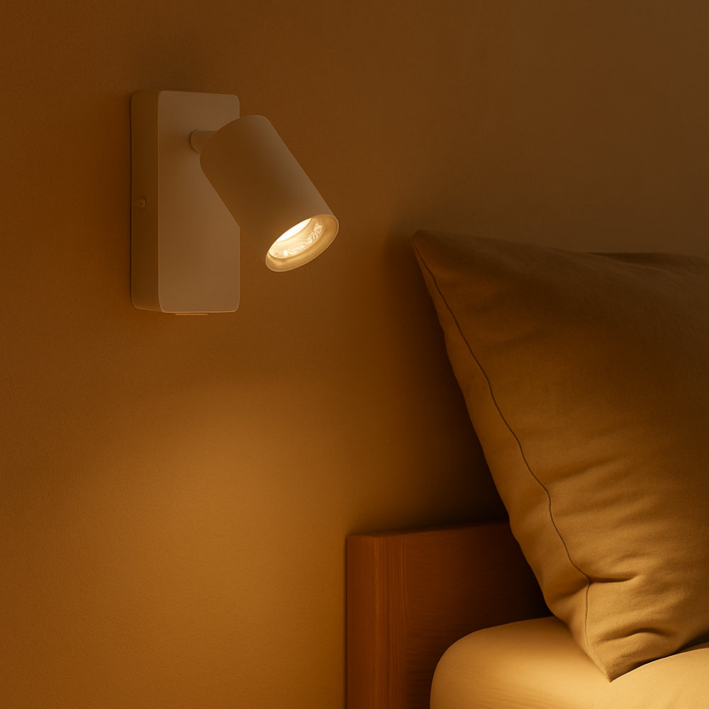 Wandlamp in moderne witte afwerking, gemonteerd boven een bed als sfeervolle leesverlichting – zijaanzicht in warme verlichting.
