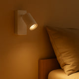 Wandlamp in moderne witte afwerking, gemonteerd boven een bed als sfeervolle leesverlichting – zijaanzicht in warme verlichting.