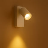 Wandspot in moderne witte uitvoering met verstelbare LED-lamp, schuin van voren gefotografeerd tegen een warme, beige muur met warm wit licht gericht naar beneden.
