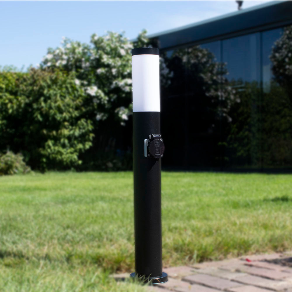 Zwarte tuinlamp met stopcontact in cilindervormig design, vooraanzicht, geplaatst op een grasveld bij een modern huis met glazen gevel.