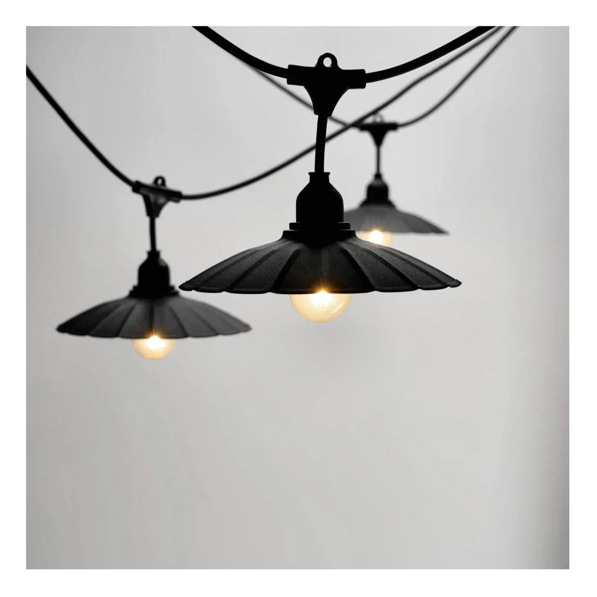 Close-up zijaanzicht van een zwarte outdoor lichtslinger met drie brandende lampen, voorzien van vintage stijl metalen kappen tegen een neutrale achtergrond.