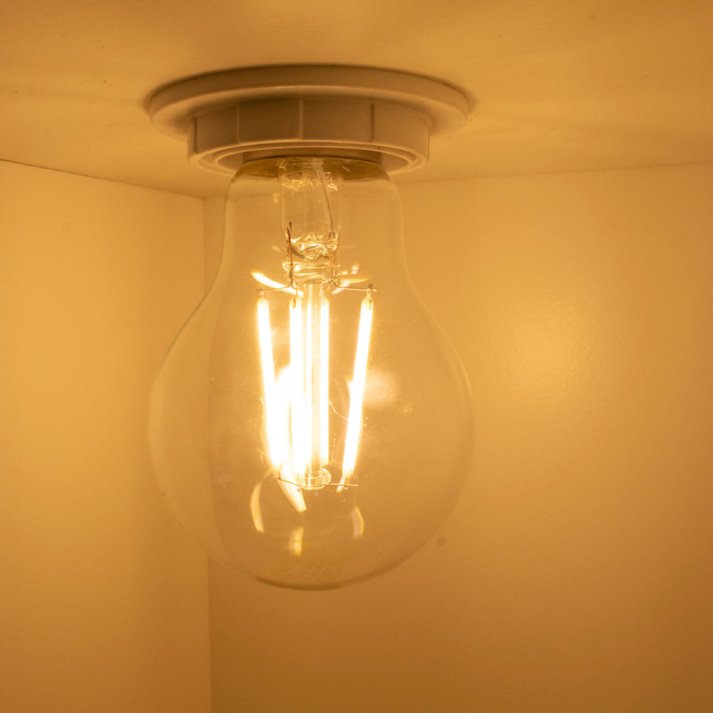 Close-up van een brandende LED filamentlamp in plafondfitting, warm wit licht uitstralend tegen een beige achtergrond, onderaanzicht.