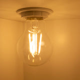 Close-up van een brandende LED filamentlamp in plafondfitting, warm wit licht uitstralend tegen een beige achtergrond, onderaanzicht.