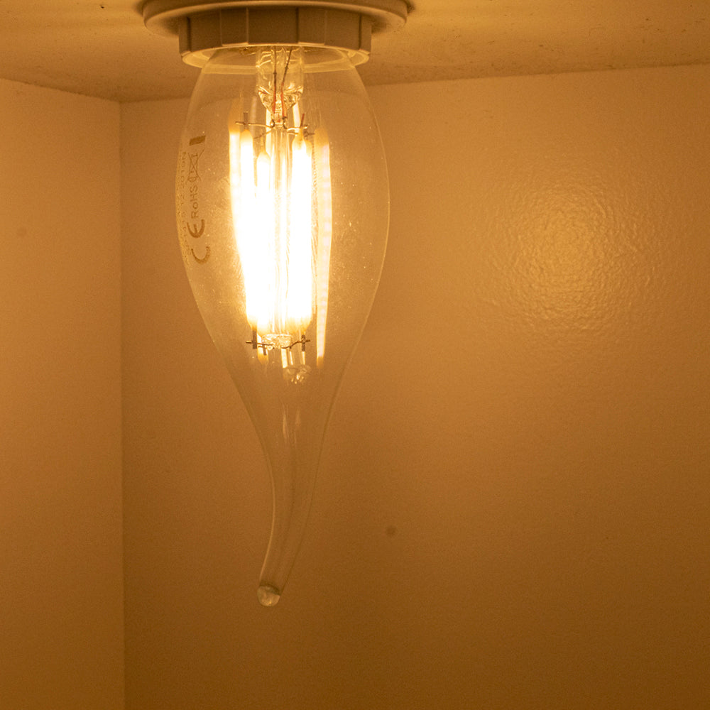 Close-up van een brandende LED filamentlamp met druppelvormig glas, gemonteerd aan het plafond, gezien vanuit een schuine onderaanzicht.