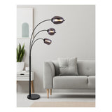 Moderne zwarte booglamp met drie gebogen armen en rookglas lampenkappen, geplaatst in een stijlvolle woonkamer naast een grijze bank – zijaanzicht.