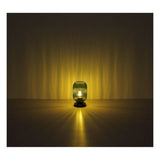 Sfeervolle frontale afbeelding van de Blomus lantaarn Ani in olijfgroen, geplaatst op een tafel in een donkere ruimte met warme lichtstralen die zich rondom verspreiden.