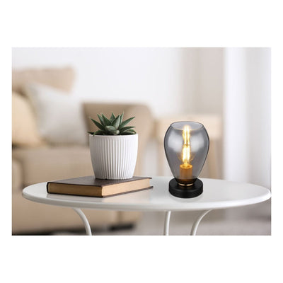 Decoratieve tafellamp met smoke-glas en retro Edison LED-lamp, geplaatst op een witte ovale salontafel naast een boek en een kleine kamerplant in een ribbelige witte pot, gefotografeerd in vooraanzicht in een modern interieur.