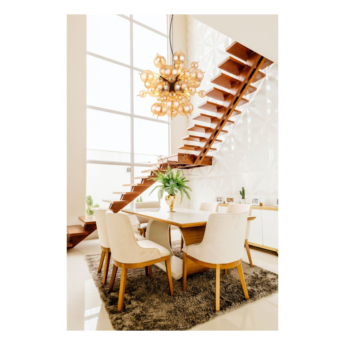 Moderne eettafelset met zes beige stoffen stoelen en een houten eettafel op een grijze vloerkleed, gezien vanuit een frontaal aanzicht in een lichte eetkamer met hoge plafonds, zwevende houten trap, grote ramen en een opvallende gouden bolvormige design hanglamp.