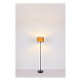 Staande vloerlamp met ronde zwarte metalen voet en industriële draadlampenkap met warmgele binnenbekleding, gefotografeerd van voren tegen een neutrale witte achtergrond.