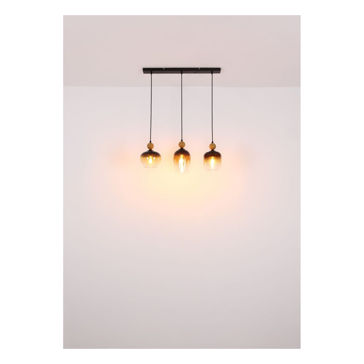 Hanglamp met drie amberkleurige glazen kappen in industriële stijl, gezien vanaf de voorzijde gemonteerd aan het plafond, met warm licht dat tegen een witte muur schijnt.