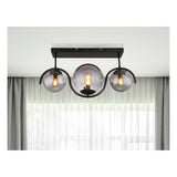 Moderne zwarte plafondlamp met drie glazen bollen en zichtbaar filament, frontaal aanzicht tegen een witte plafondachtergrond met gordijnen en natuurlijk licht.