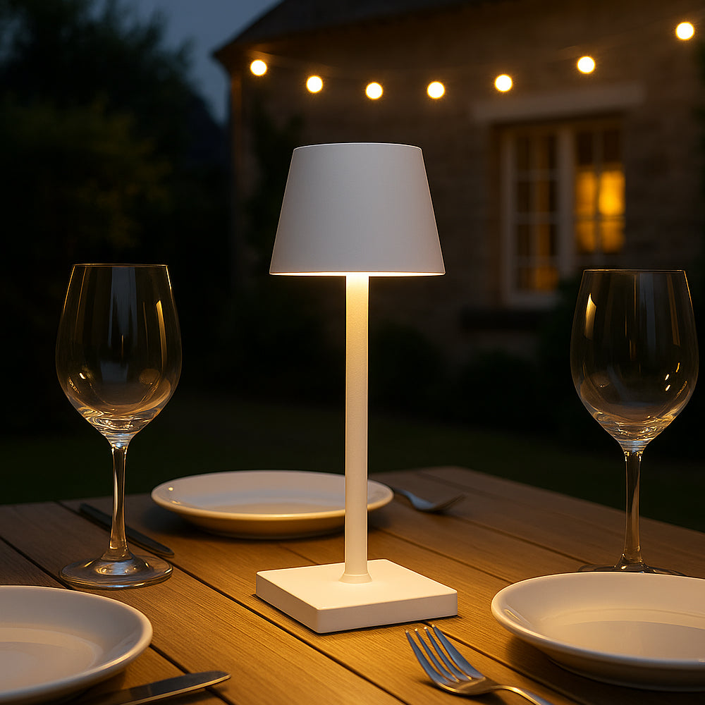 Moderne witte tafellamp op houten buitentafel tijdens schemering, omringd door borden en wijnglazen, sfeervol verlicht met achtergrond van tuinverlichting en woning – frontaal aanzicht.