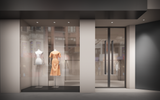 Etalageaanzicht van een moderne kledingwinkel met twee paspoppen, waarvan één gekleed is in een elegante perzikkleurige jurk, links in de vitrine; rechts een glazen winkeldeur met zicht op een minimalistisch interieur en een wit kledingstuk aan een hanger.
