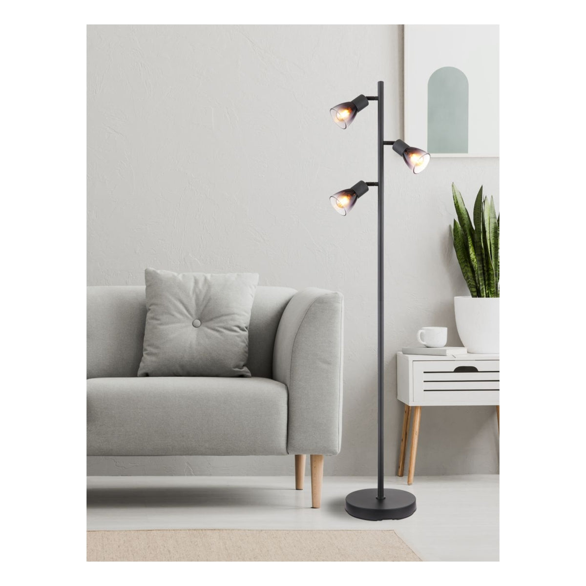 Moderne zwarte vloerlamp met drie verstelbare spots, geplaatst in een minimalistisch interieur met een grijze bank, witte bijzettafel en een kamerplant – frontaal aanzicht.