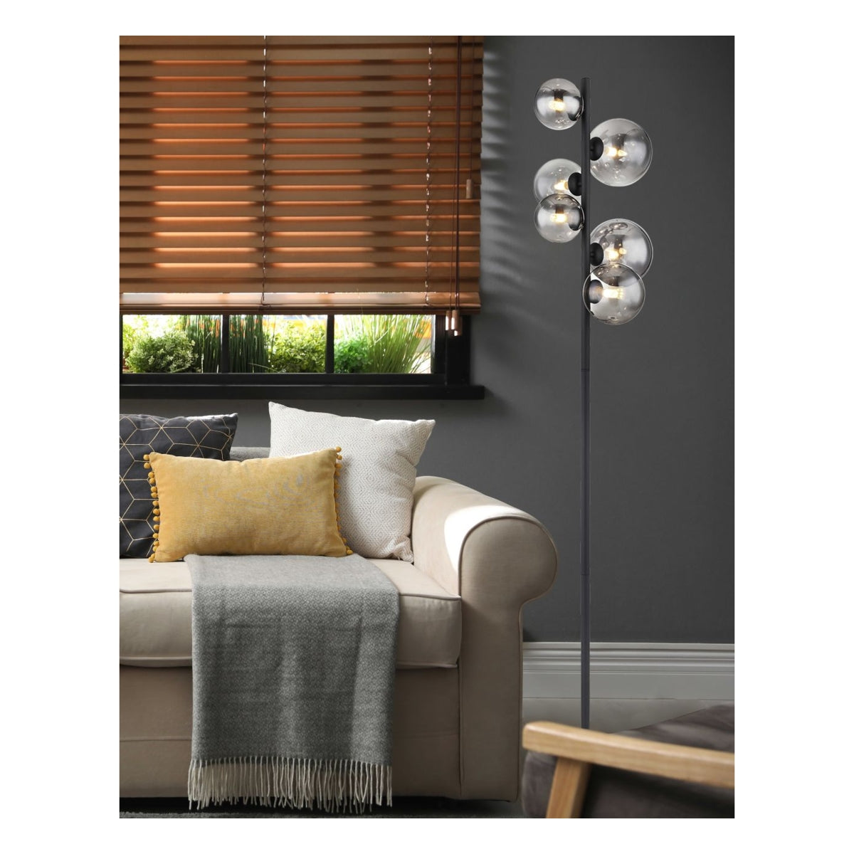 Moderne staande lamp met zes ronde glazen bollen op zwarte metalen standaard, geplaatst naast een beige bank in een stijlvolle woonkamer – zijaanzicht met focus op het product in zijn interieurcontext.