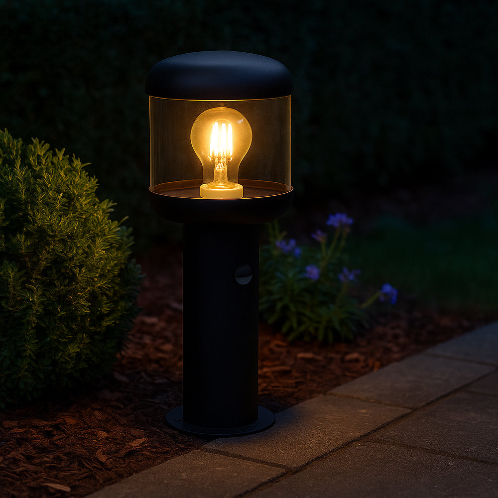 Moderne zwarte buitenlamp met amberkleurige LED-lichtbron, staand op een tuinpad tussen groen struikgewas, gefotografeerd in schemering van voren met focus op warm licht en stijlvolle cilindervormige behuizing.