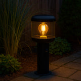 Zwart stalen buitenlamp op sokkel met filament LED-lamp, zijkantaanzicht bij schemering in een tuin met siergrassen en bestrating.