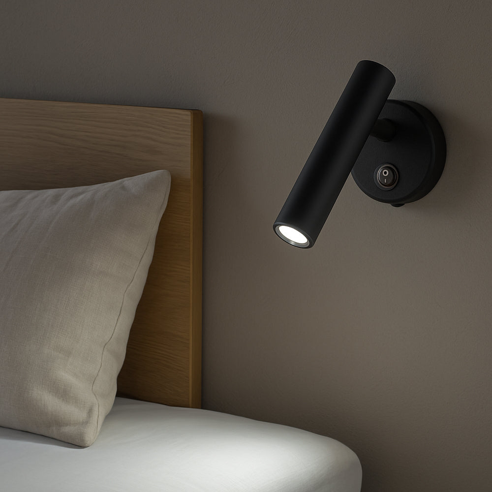 Zwarte moderne wandlamp met cilindervormig design, gemonteerd naast een houten bedframe en gericht op het bedoppervlak; zijaanzicht van de lamp in gebruik als leeslampje bij het bed.