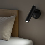 Zwarte moderne wandlamp met cilindervormig design, gemonteerd naast een houten bedframe en gericht op het bedoppervlak; zijaanzicht van de lamp in gebruik als leeslampje bij het bed.