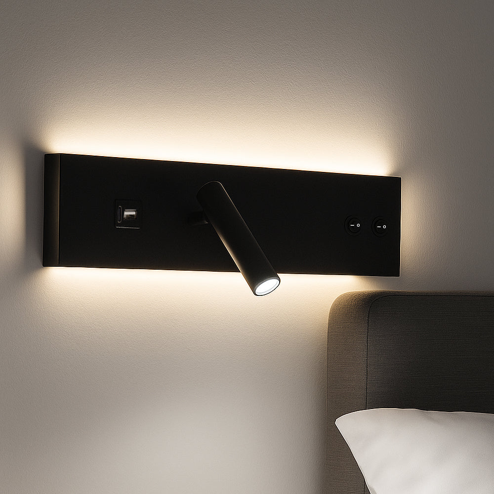Zijaanzicht van een moderne zwarte wandlamp met geïntegreerd leeslampje, USB-poort en indirecte achtergrondverlichting, gemonteerd boven een bed tegen een lichtgekleurde muur.