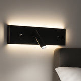 Zijaanzicht van een moderne zwarte wandlamp met geïntegreerd leeslampje, USB-poort en indirecte achtergrondverlichting, gemonteerd boven een bed tegen een lichtgekleurde muur.