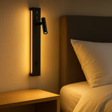 Moderne zwarte wandlamp met geïntegreerde LED-verlichting en leeslampje, gemonteerd naast een bed met houten nachtkastje en stoffen hoofdbord, vooraanzicht in sfeervolle slaapkamerverlichting.