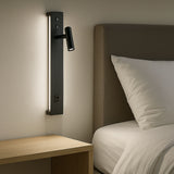 Moderne zwarte wandlamp met geïntegreerd leeslampje en sfeerverlichting, gemonteerd naast een bed met houten nachtkastje – zijaanzicht in een slaapkameromgeving.