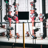 Industriële LED-bouwlamp op statief, achteraanzicht, geplaatst in een technische ruimte met leidingen en rode afsluiters op de achtergrond.