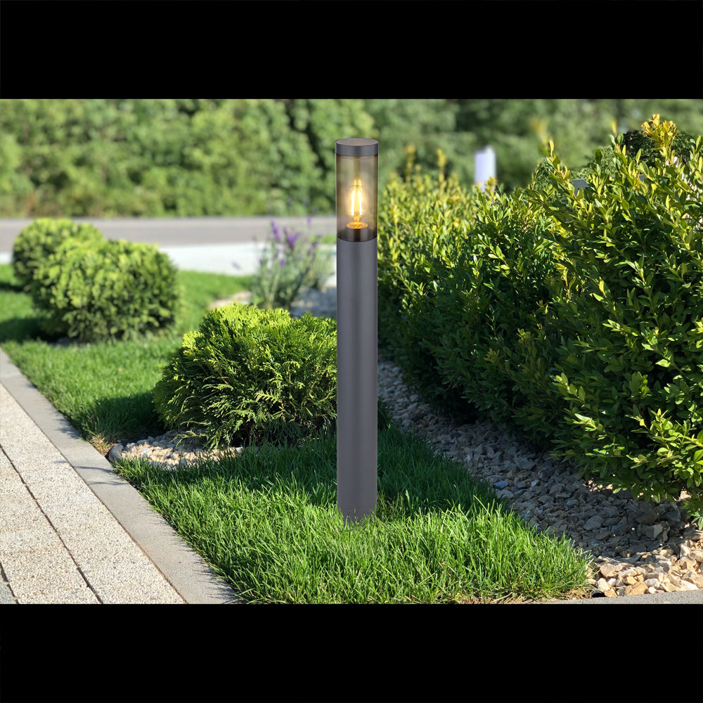 Moderne zwarte tuinlamp op zonne-energie met zichtbaar filament, staand in een groene tuin langs een borderpad – frontaal aanzicht bij daglicht.