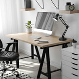 Moderne thuiswerkplek met een licht houten bureau met zwarte schragen, voorzien van een computer, bureaulamp, notitieboekje en plant, gefotografeerd in een zijaanzicht met daglichtinval vanaf links.