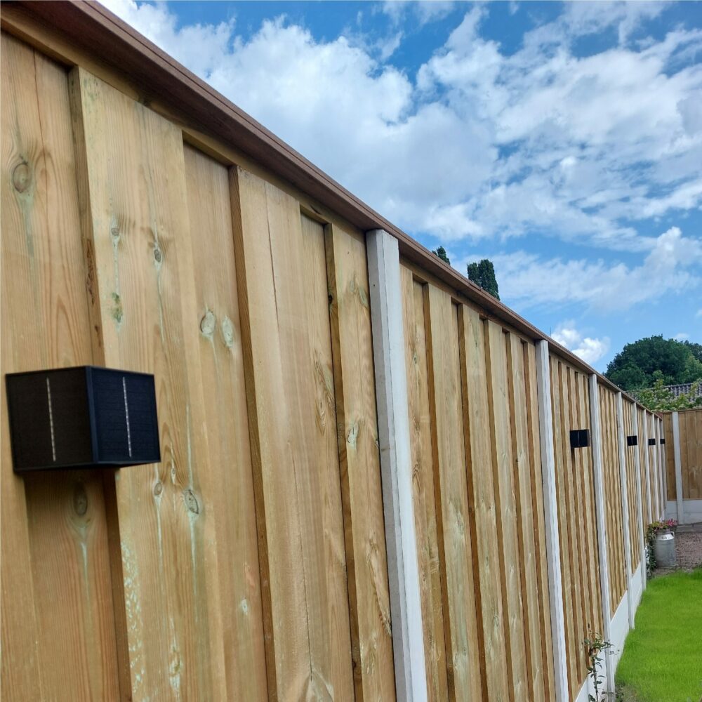 Houten schutting met betonpalen en geïntegreerde zwarte solar wandlampen, zijaanzicht op een zonnige dag met blauwe lucht en graspartij op de voorgrond.