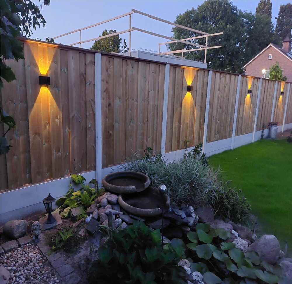 Houten schutting met verticale planken voorzien van moderne wandlampen met warm wit licht, tuinzicht bij schemering met sierlijke beplanting, vijver en grasveld – zijaanzicht van de tuin en schutting.