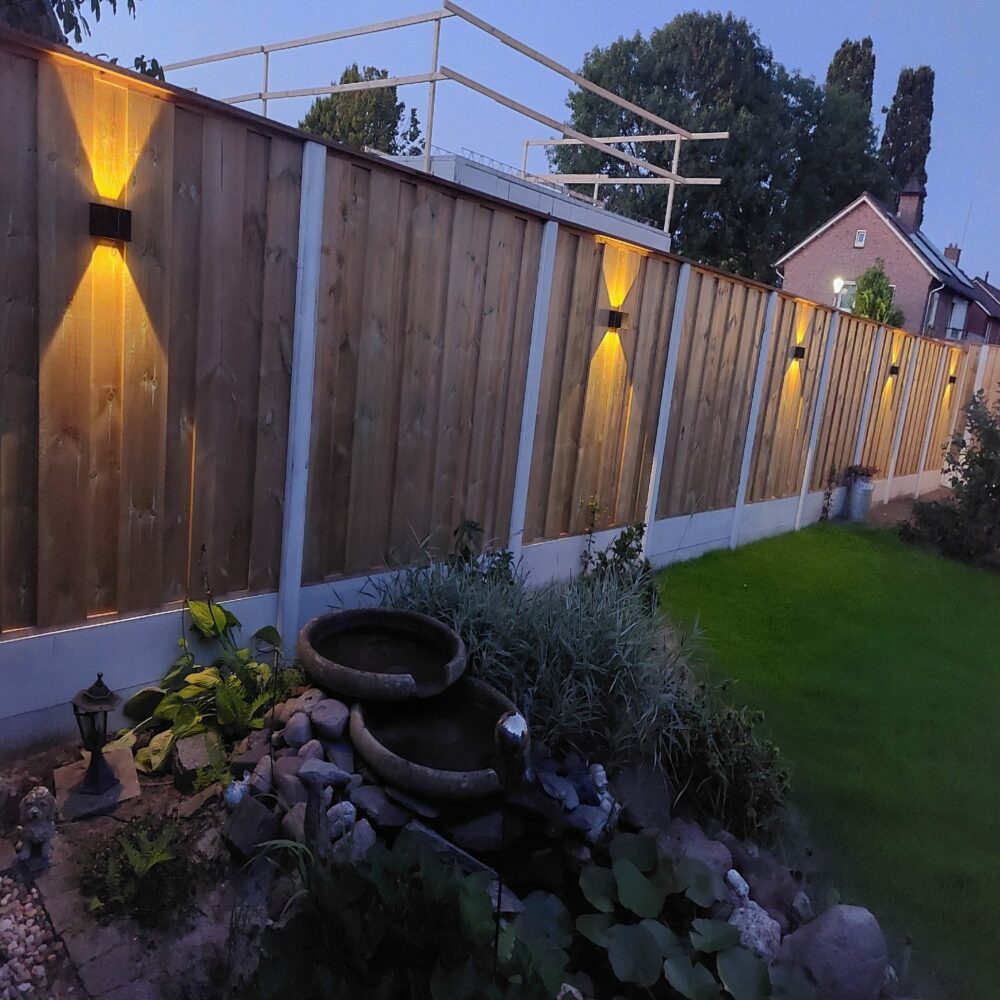 Houten schutting met geïntegreerde wandverlichting in schemerlicht, zij-aanzicht van de tuin met spotlight-effect op de planken en zicht op een kleine vijver met stapelstenen en beplanting op de voorgrond.