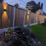 Houten schutting met geïntegreerde wandverlichting in schemerlicht, zij-aanzicht van de tuin met spotlight-effect op de planken en zicht op een kleine vijver met stapelstenen en beplanting op de voorgrond.