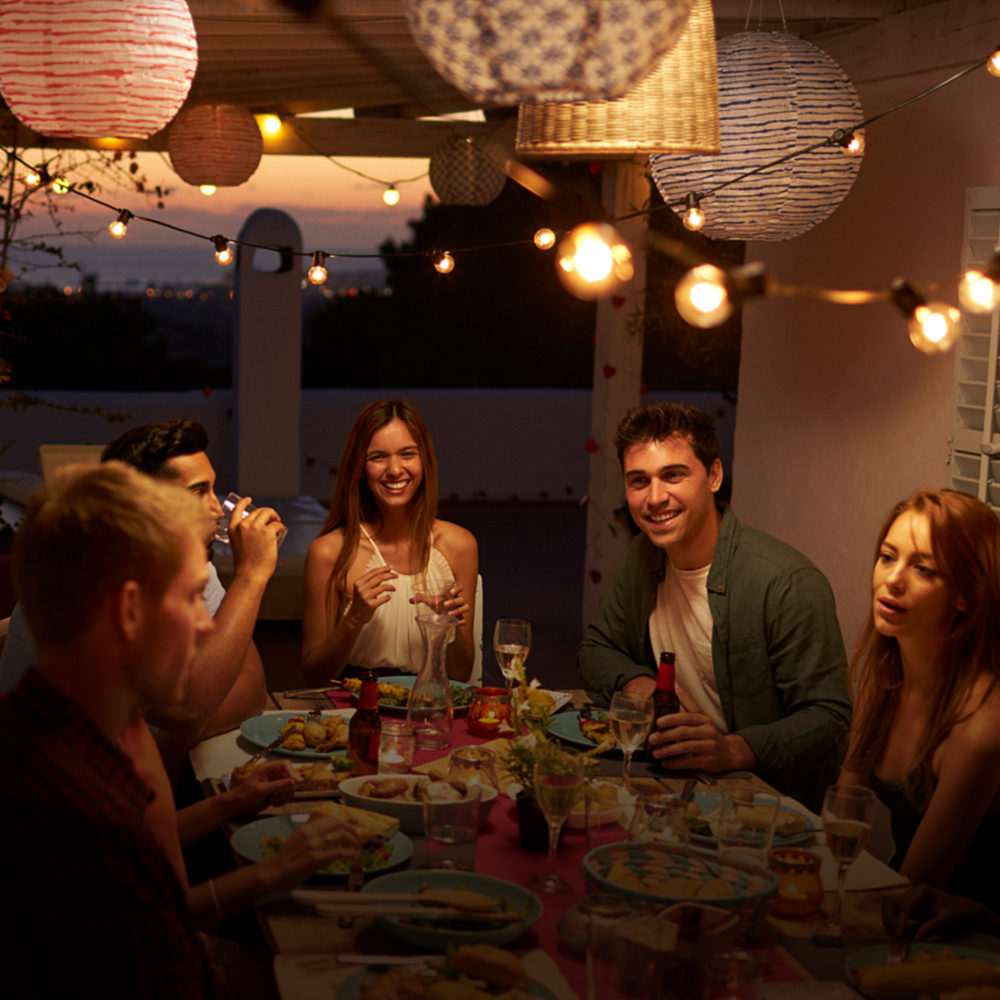 Groep vrienden geniet van een gezellige dineravond onder een overdekt terras bij zonsondergang, met sfeervolle buitenverlichting en kleurrijke lantaarns – vooraanzicht van het tafereel.