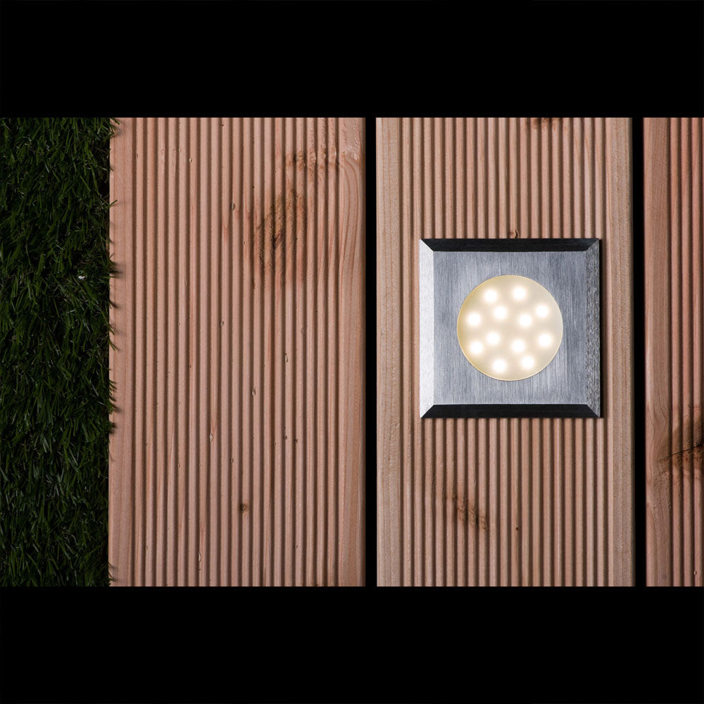 Vierkante LED inbouwspot voor buitenverlichting, gemonteerd in een houten terrasvloer naast kunstgras, bovenaanzicht met ingeschakelde verlichting.