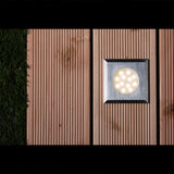 Vierkante LED inbouwspot voor buitenverlichting, gemonteerd in een houten terrasvloer naast kunstgras, bovenaanzicht met ingeschakelde verlichting.