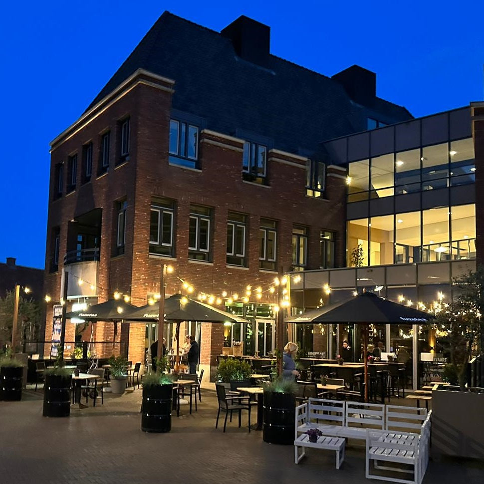Avondaanzicht van een modern stadsgebouw met een sfeervol verlicht terras van een restaurant, voorzien van parasols, tafels en stoelen in een gezellige buitenomgeving.