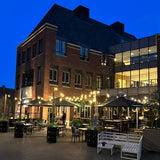Avondaanzicht van een sfeervol verlicht terras met zwarte parasols en buitenzitplaatsen voor een modern bakstenen gebouw met grote ramen en een glazen gevel, ideaal voor horeca of restaurantpromotie.