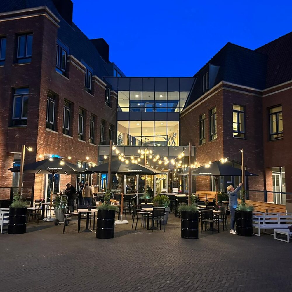 Avondaanzicht van het terras van restaurant Remise56, sfeervol verlicht met stringverlichting en omgeven door een historisch bakstenen gebouw met moderne glazen entree.