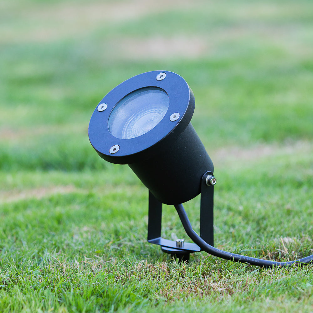 Zwarte buitenspot met ronde kap en grondpin, schuin van voren gefotografeerd op een gazon, geschikt voor tuinverlichting of het aanlichten van objecten in de buitenruimte.