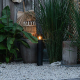 Zwarte moderne buitenlamp sokkelverlichting met zichtbare filament LED-lamp, frontaal aanzicht geplaatst tussen siergrassen en decoratieve elementen op een grindondergrond in een sfeervolle tuinsetting.