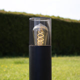 Moderne zwarte buitenlamp met zichtbare filament LED-lamp, cilindervormig ontwerp met glazen kap, vooraanzicht tegen groene tuinachtergrond.