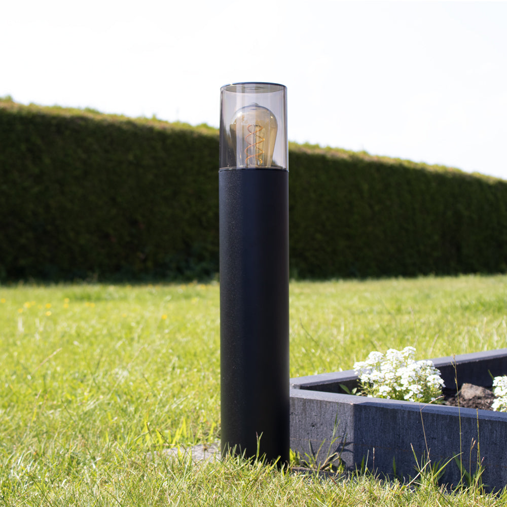 Moderne zwarte buitenlamp sokkellamp met E27 fitting, cilindervormig ontwerp en transparante bovenkap met zichtbare filamentlamp, gefotografeerd in zijaanzicht op een zonnige dag in een tuin.