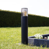 Moderne zwarte buitenlamp sokkellamp met E27 fitting, cilindervormig ontwerp en transparante bovenkap met zichtbare filamentlamp, gefotografeerd in zijaanzicht op een zonnige dag in een tuin.