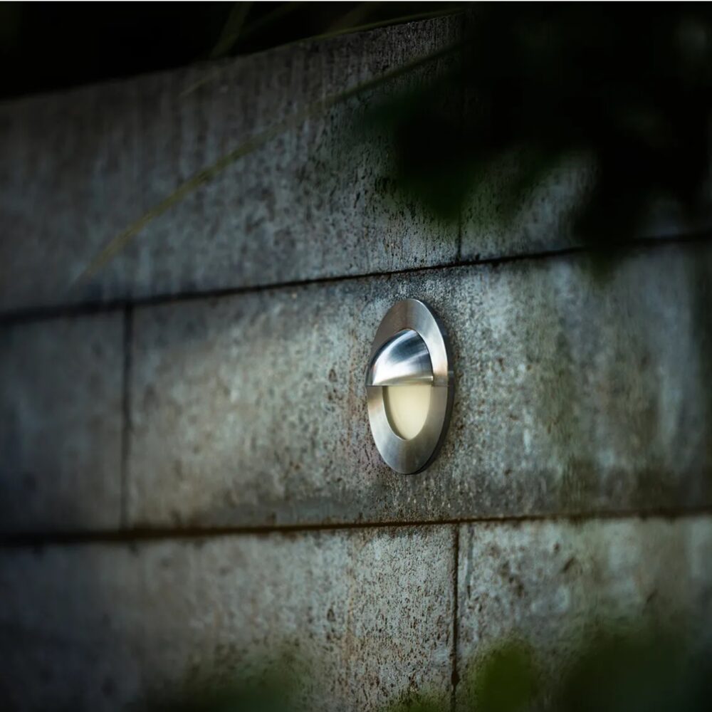 Ronde roestvrijstalen wandlamp gemonteerd op een betonnen muur, weergegeven in een diagonaal zijaanzicht bij schemerlicht, ideaal voor buitenverlichting of tuinpadverlichting.