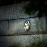 Ronde roestvrijstalen wandlamp gemonteerd op een betonnen muur, weergegeven in een diagonaal zijaanzicht bij schemerlicht, ideaal voor buitenverlichting of tuinpadverlichting.