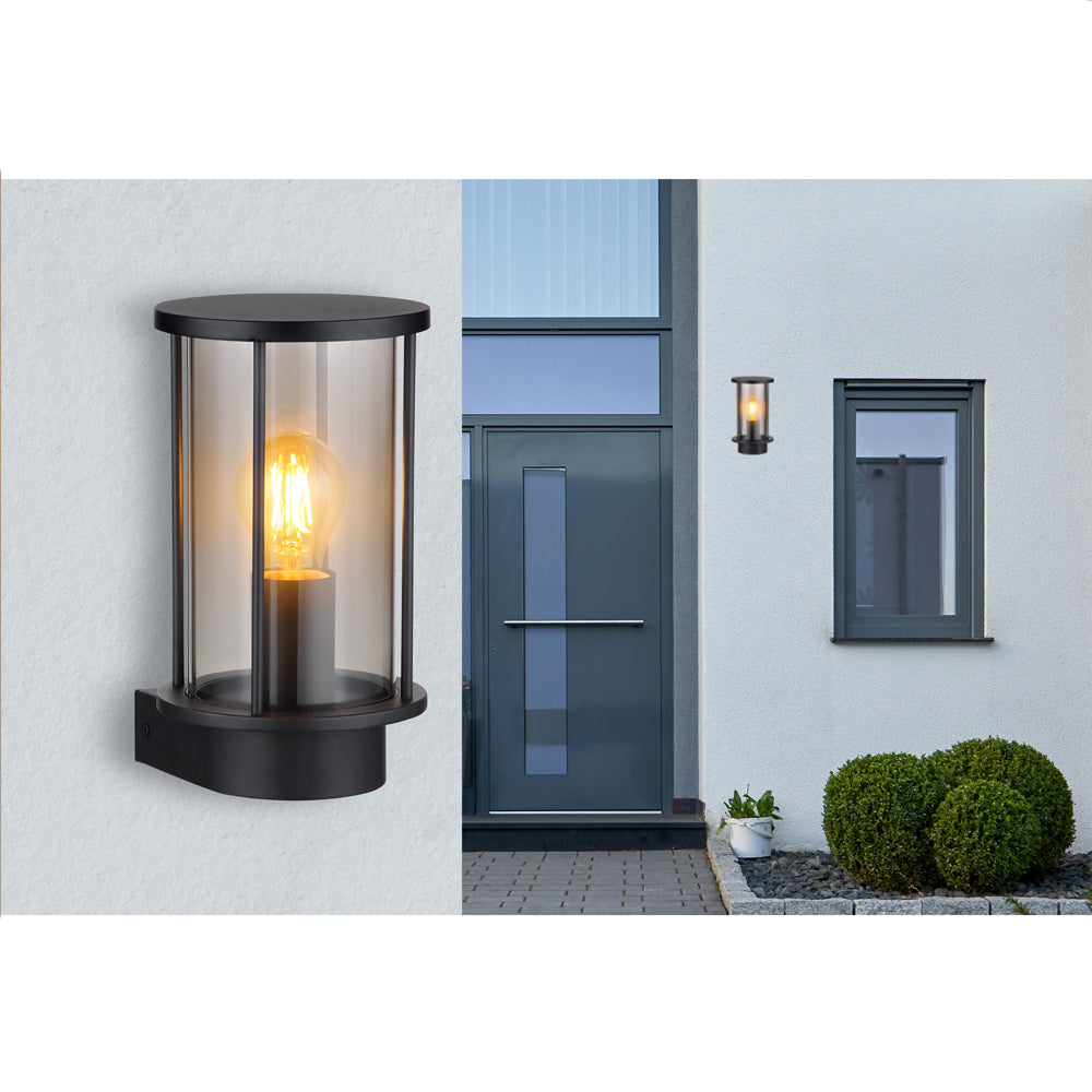 Moderne zwarte cilindervormige buitenwandlamp met glazen behuizing en zichtbare filamentlamp, getoond in close-up en gemonteerd naast een voordeur aan de gevel van een woning – vooraanzicht.