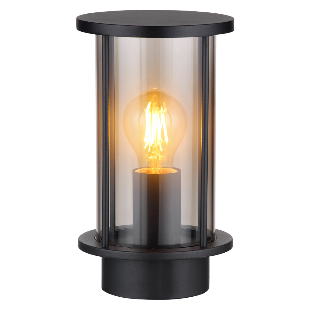 Moderne zwarte buitenlamp met cilindervormig design en heldere glazen behuizing, zichtbaar van schuin vooraanzicht; voorzien van een warm wit brandende filament LED-lamp.