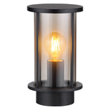 Moderne zwarte buitenlamp met cilindervormig design en heldere glazen behuizing, zichtbaar van schuin vooraanzicht; voorzien van een warm wit brandende filament LED-lamp.